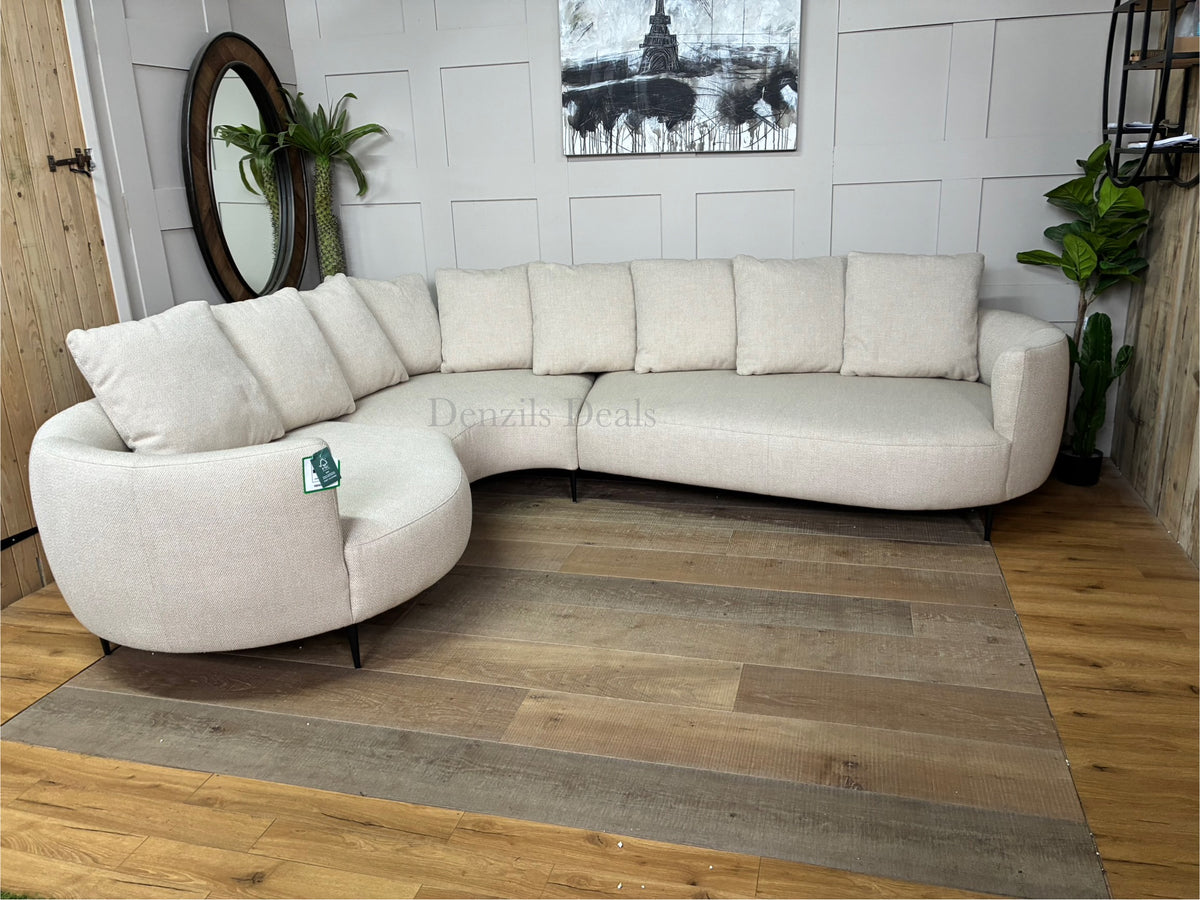 A contemporary 5-seater corner sofa by John Lewis. The sofa has a distinctive fluid design with soft, rounded organic edges. It is upholstered in a heavily textured, light beige 'twisted boucle' fabric in the shade 'Putty'. The unit curves into a left-hand facing configuration, supported by low-profile slim metal legs, and is topped with multiple matching square pillow cushions