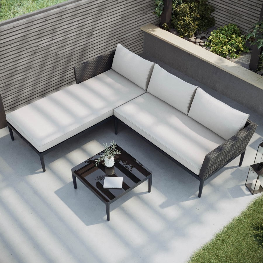 A 4-seater black PE rattan outdoor corner sofa with an extended chaise section, light beige thick cushions, and a low square coffee table with a tempered glass top.