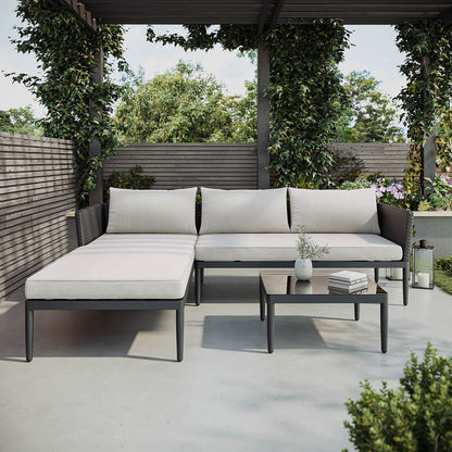 A 4-seater black PE rattan outdoor corner sofa with an extended chaise section, light beige thick cushions, and a low square coffee table with a tempered glass top.