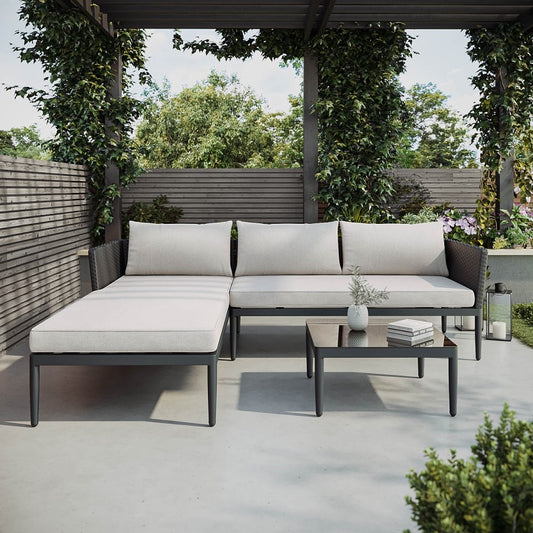 A 4-seater black PE rattan outdoor corner sofa with an extended chaise section, light beige thick cushions, and a low square coffee table with a tempered glass top.
