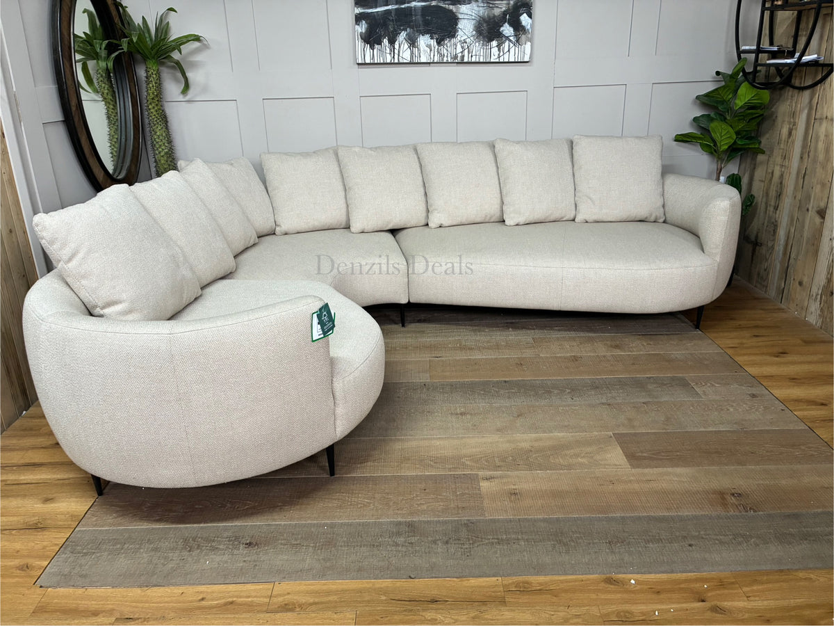 A contemporary 5-seater corner sofa by John Lewis. The sofa has a distinctive fluid design with soft, rounded organic edges. It is upholstered in a heavily textured, light beige 'twisted boucle' fabric in the shade 'Putty'. The unit curves into a left-hand facing configuration, supported by low-profile slim metal legs, and is topped with multiple matching square pillow cushions