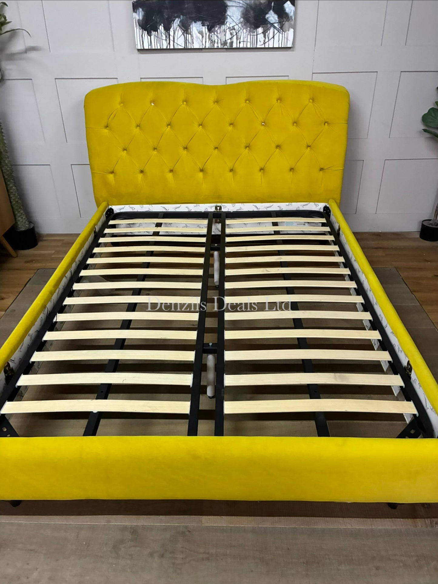 A brightly lit bedroom featuring the Joules Windsor king-size bed frame from DFS in mustard yellow velvet. The bed is dressed in neutral linen sheets, showcasing its plush, deeply buttoned Chesterfield headboard and unique colour-dipped wooden feet against a modern backdrop