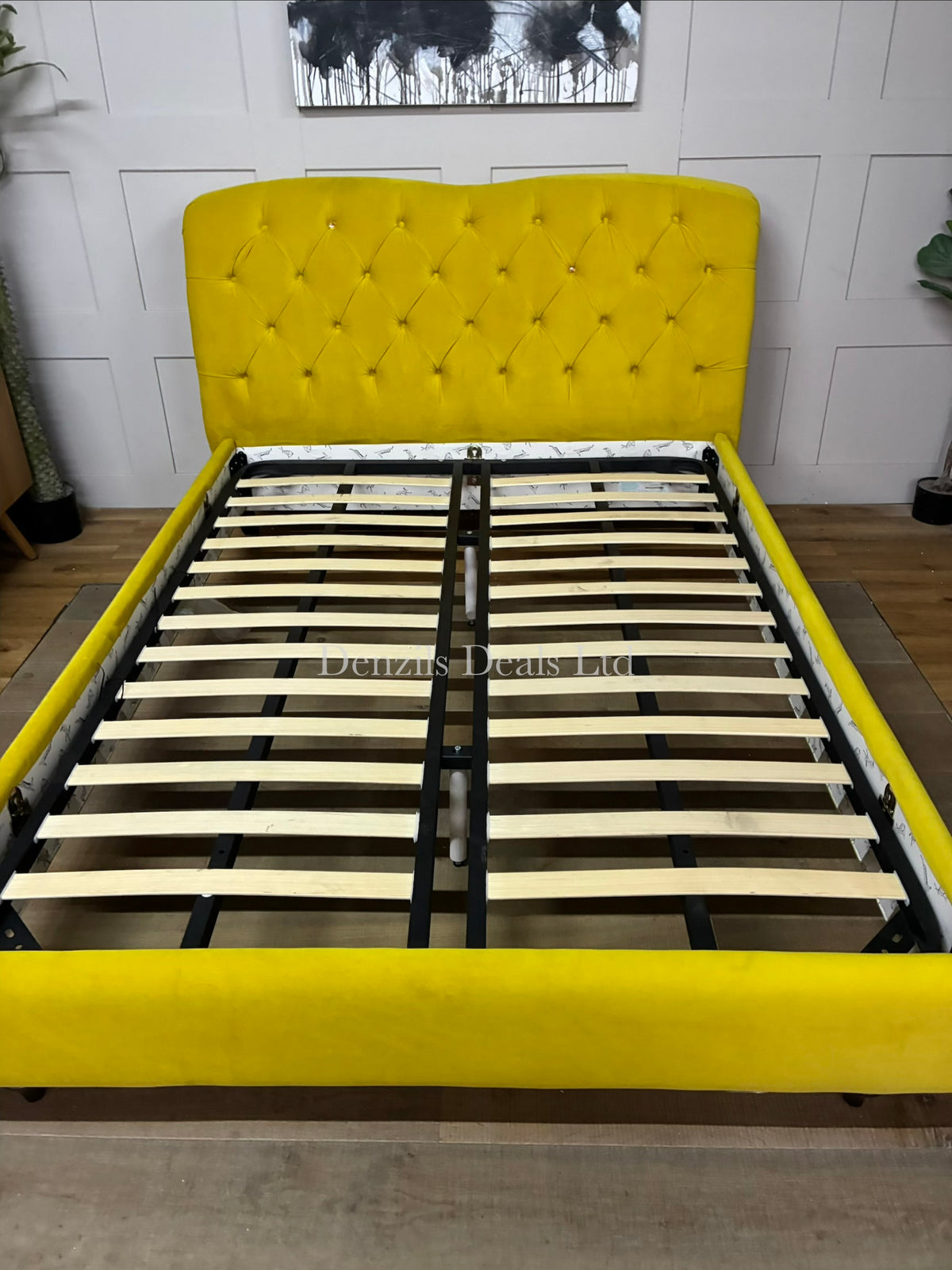 A brightly lit bedroom featuring the Joules Windsor king-size bed frame from DFS in mustard yellow velvet. The bed is dressed in neutral linen sheets, showcasing its plush, deeply buttoned Chesterfield headboard and unique colour-dipped wooden feet against a modern backdrop