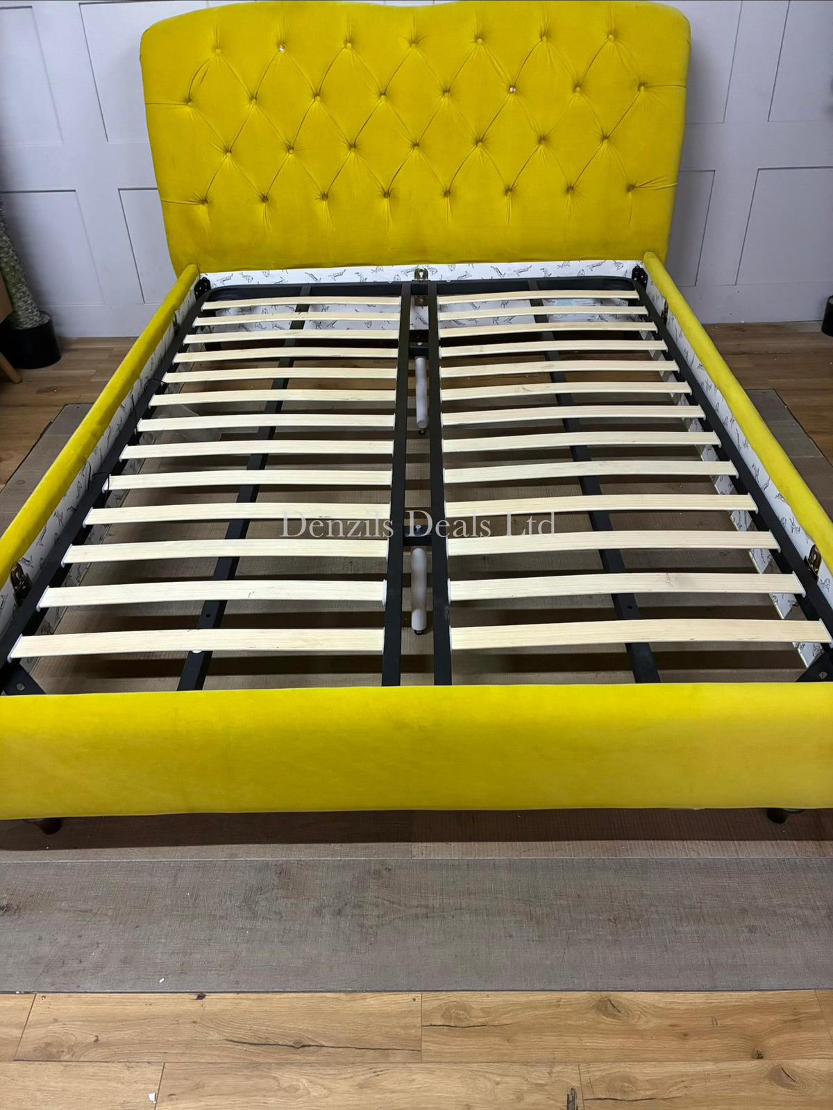 A brightly lit bedroom featuring the Joules Windsor king-size bed frame from DFS in mustard yellow velvet. The bed is dressed in neutral linen sheets, showcasing its plush, deeply buttoned Chesterfield headboard and unique colour-dipped wooden feet against a modern backdrop