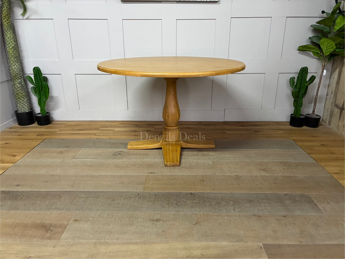 A circular Oak Furnitureland Henley dining table crafted from light natural oak wood, showcasing a smooth round tabletop and a prominent, farmhouse-style carved central pedestal leg.