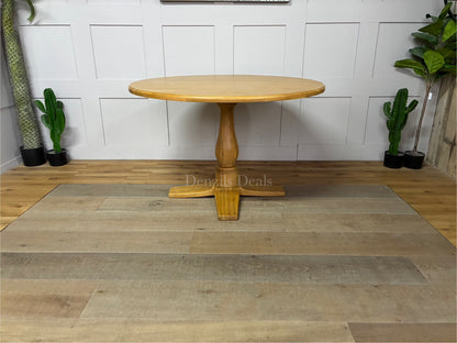 A circular Oak Furnitureland Henley dining table crafted from light natural oak wood, showcasing a smooth round tabletop and a prominent, farmhouse-style carved central pedestal leg.