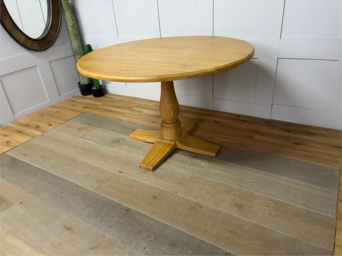 A circular Oak Furnitureland Henley dining table crafted from light natural oak wood, showcasing a smooth round tabletop and a prominent, farmhouse-style carved central pedestal leg."