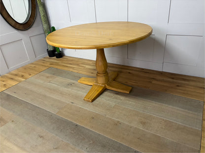 A circular Oak Furnitureland Henley dining table crafted from light natural oak wood, showcasing a smooth round tabletop and a prominent, farmhouse-style carved central pedestal leg."