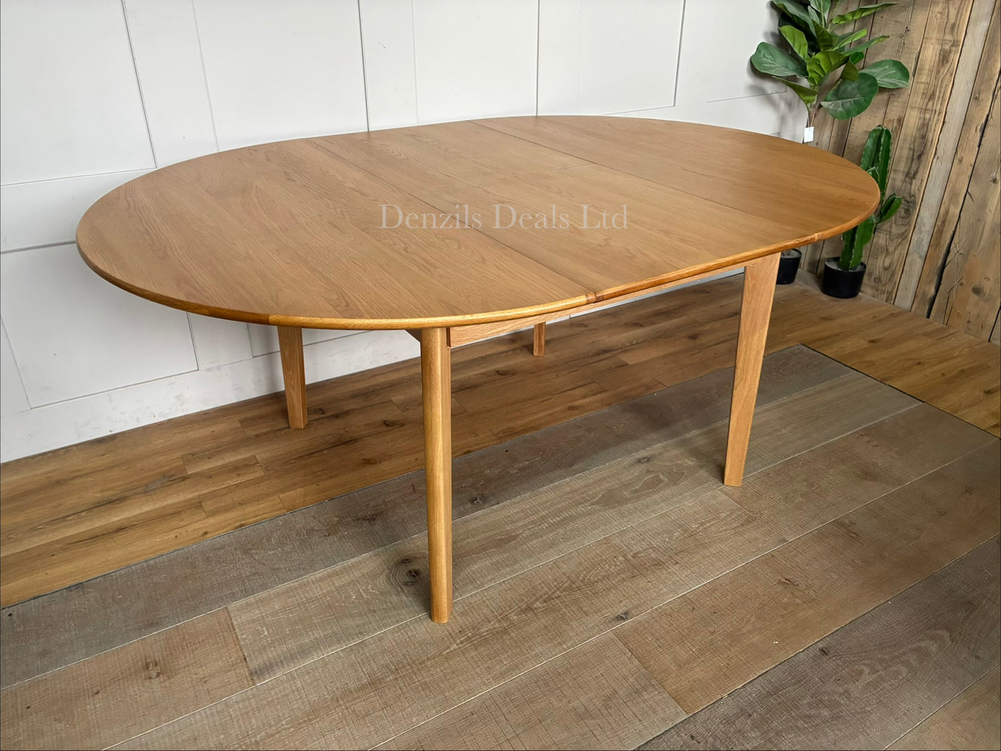 A circular light oak dining table featuring an integrated butterfly extension leaf that expands the tabletop into an oval shape.