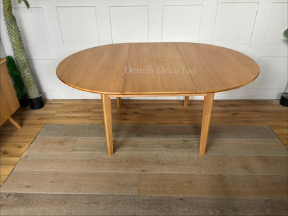 A circular light oak dining table featuring an integrated butterfly extension leaf that expands the tabletop into an oval shape.