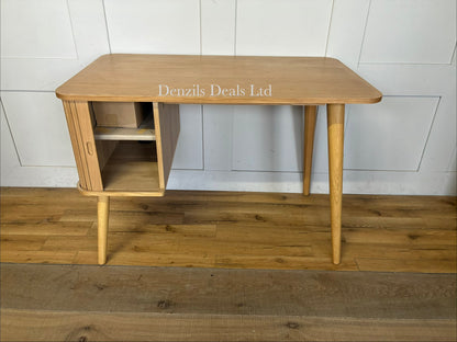 A compact Scandi-style wooden writing desk with tapered legs, a rectangular work surface, and a cabinet section featuring a sliding slatted door on one side."