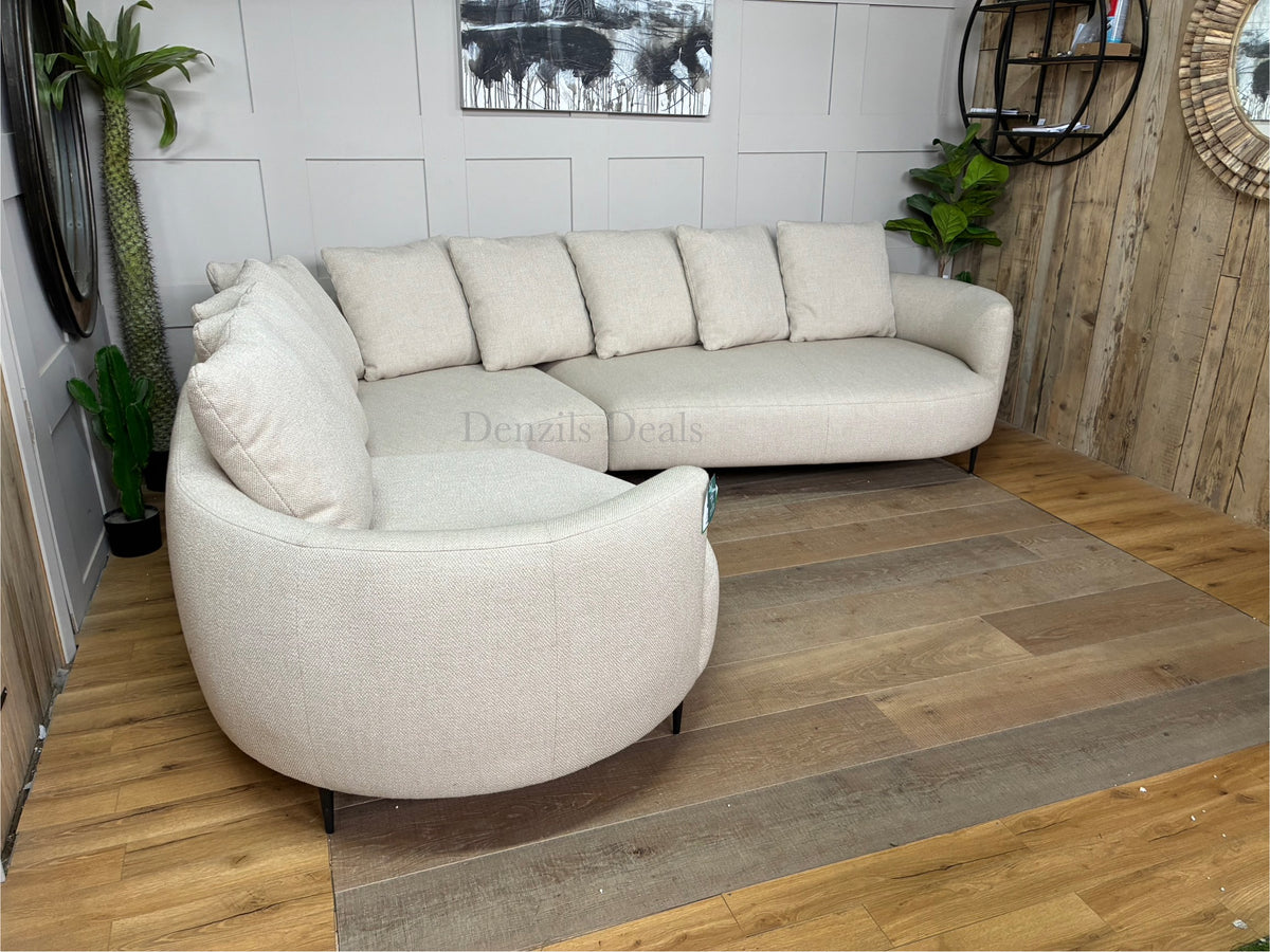 A contemporary 5-seater corner sofa by John Lewis. The sofa has a distinctive fluid design with soft, rounded organic edges. It is upholstered in a heavily textured, light beige 'twisted boucle' fabric in the shade 'Putty'. The unit curves into a left-hand facing configuration, supported by low-profile slim metal legs, and is topped with multiple matching square pillow cushions