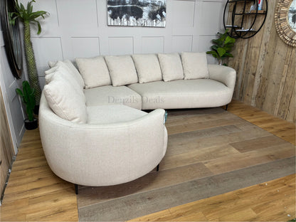 A contemporary 5-seater corner sofa by John Lewis. The sofa has a distinctive fluid design with soft, rounded organic edges. It is upholstered in a heavily textured, light beige 'twisted boucle' fabric in the shade 'Putty'. The unit curves into a left-hand facing configuration, supported by low-profile slim metal legs, and is topped with multiple matching square pillow cushions