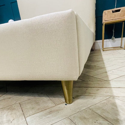 A contemporary double bed frame with a low-profile design. The headboard and frame are fully upholstered in a neutral stone-coloured fabric. It features vertical external seams on the corners and sits on four simple, angled wooden legs