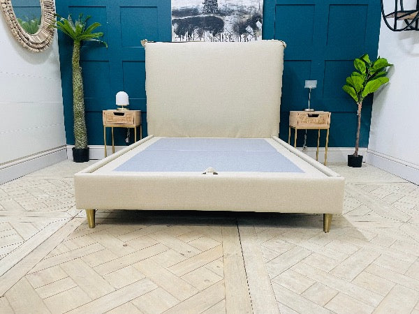 A contemporary double bed frame with a low-profile design. The headboard and frame are fully upholstered in a neutral stone-coloured fabric. It features vertical external seams on the corners and sits on four simple, angled wooden legs