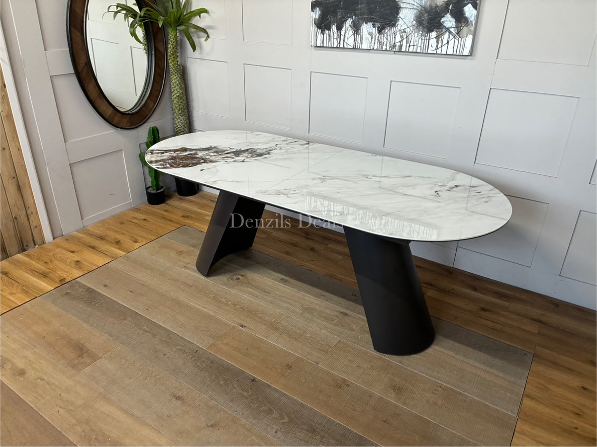 A contemporary extendable 4-to-6 seater dining table with a rounded pill-shaped top made of white ceramic with realistic grey marble veining. The table is supported by thick, angled iron legs finished in a charcoal grey color