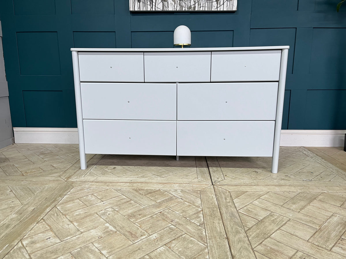 A contemporary, soft-profile John Lewis Spindle 7 Drawer Chest in a smooth grey lacquered finish. The spacious wooden chest features a combination of deep and shallow drawers arranged vertically, built with sturdy pine and MDF. It is designed with clean lines and distinctly rounded edges that soften its overall profile, offering a modern and minimalist look for a bedroom.