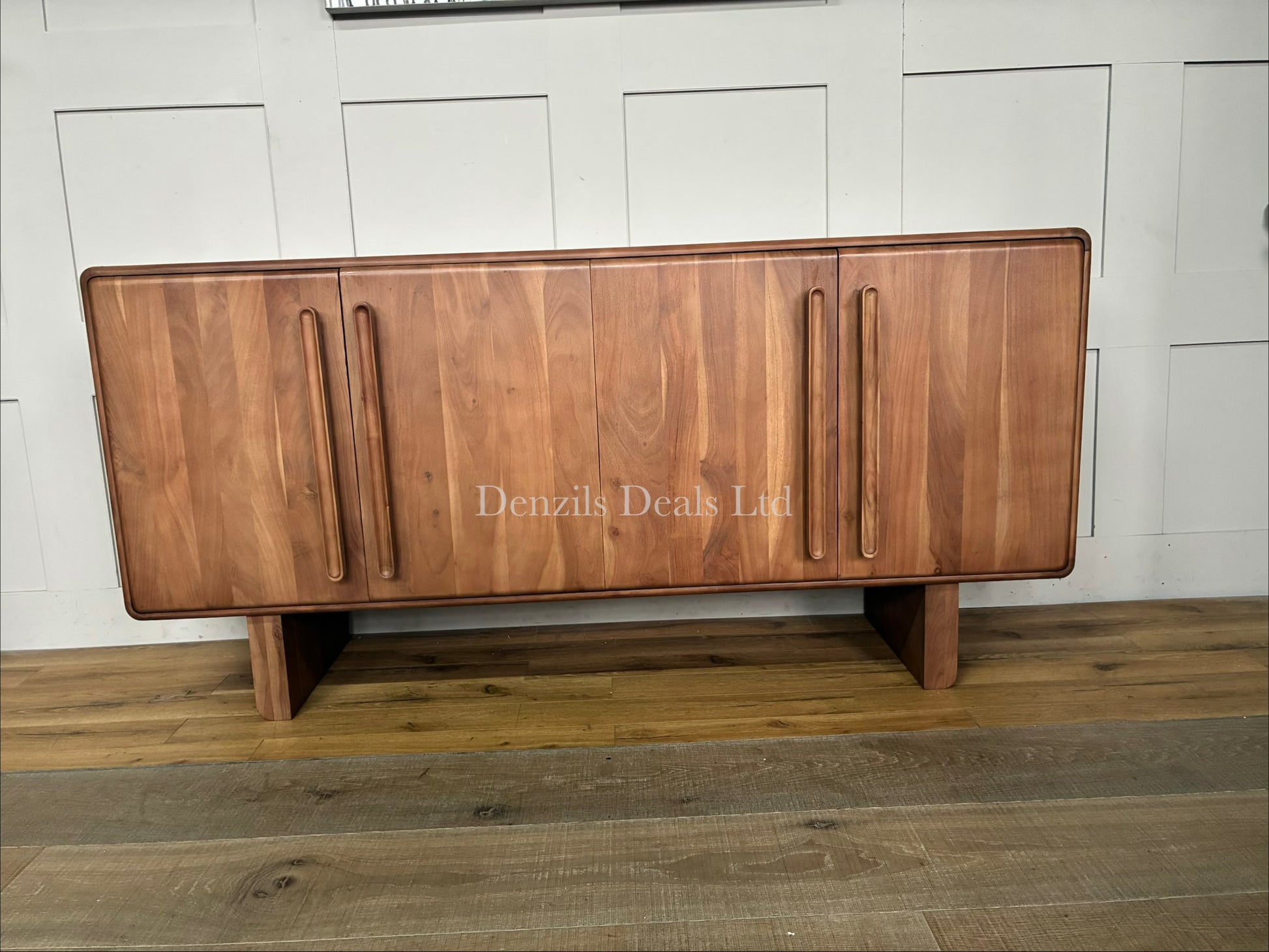 A front-facing product shot of the John Lewis Akola Sideboard in a light, natural wood tone. The sideboard is crafted from panelled acacia wood, creating vertical line patterns across its four minimalist doors. The outer edges of the frame are softly rounded, softening its blocky silhouette. The unit sits directly on the ground without legs, serving as a clean, mid-century or modern living room storage solution