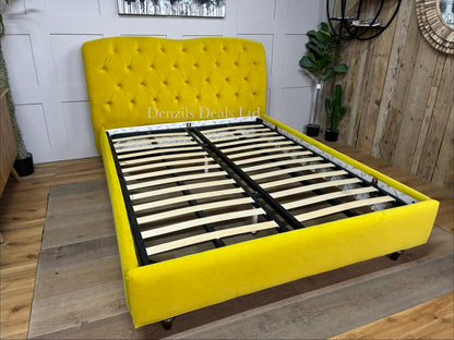 A king-size Chesterfield-style bed frame from the Joules collection at DFS, upholstered in vibrant mustard yellow velvet. The high headboard features deep button-back tufting, a rounded classic silhouette, and small hidden details like signature Joules hare motifs on the buttons. The bed frame is elevated on turned wooden feet with colour-dipped details.