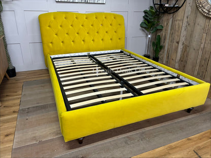 A king-size Chesterfield-style bed frame from the Joules collection at DFS, upholstered in vibrant mustard yellow velvet. The high headboard features deep button-back tufting, a rounded classic silhouette, and small hidden details like signature Joules hare motifs on the buttons. The bed frame is elevated on turned wooden feet with colour-dipped details.