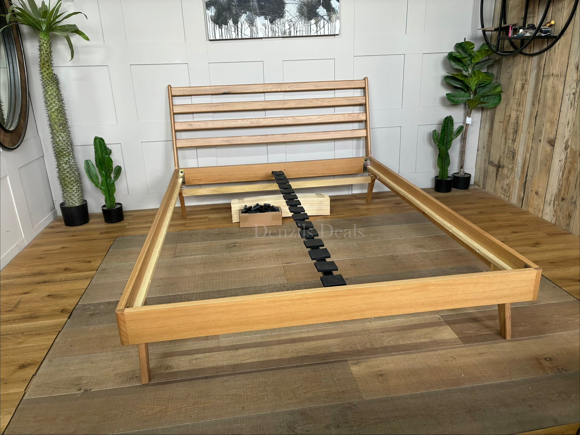 A king-size minimalist platform bed frame made of light natural oak wood. It features a high, slatted headboard with clean horizontal lines, tapered angled legs, and a sturdy sprung-slat base.