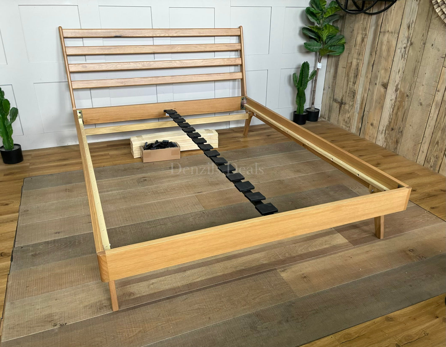 A king-size minimalist platform bed frame made of light natural oak wood. It features a high, slatted headboard with clean horizontal lines, tapered angled legs, and a sturdy sprung-slat base.