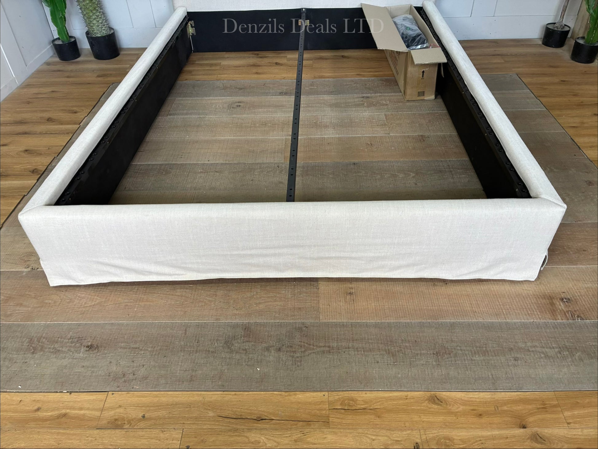 A king-size modern bedroom setup featuring the John Lewis Relax Rustic Upholstered Bed Frame. The bed has a tall, well-padded rectangular headboard covered in loose, casual-fitting natural beige fabric. The frame is built from visible timber feet at the base, supporting a low-profile matching upholstered surround