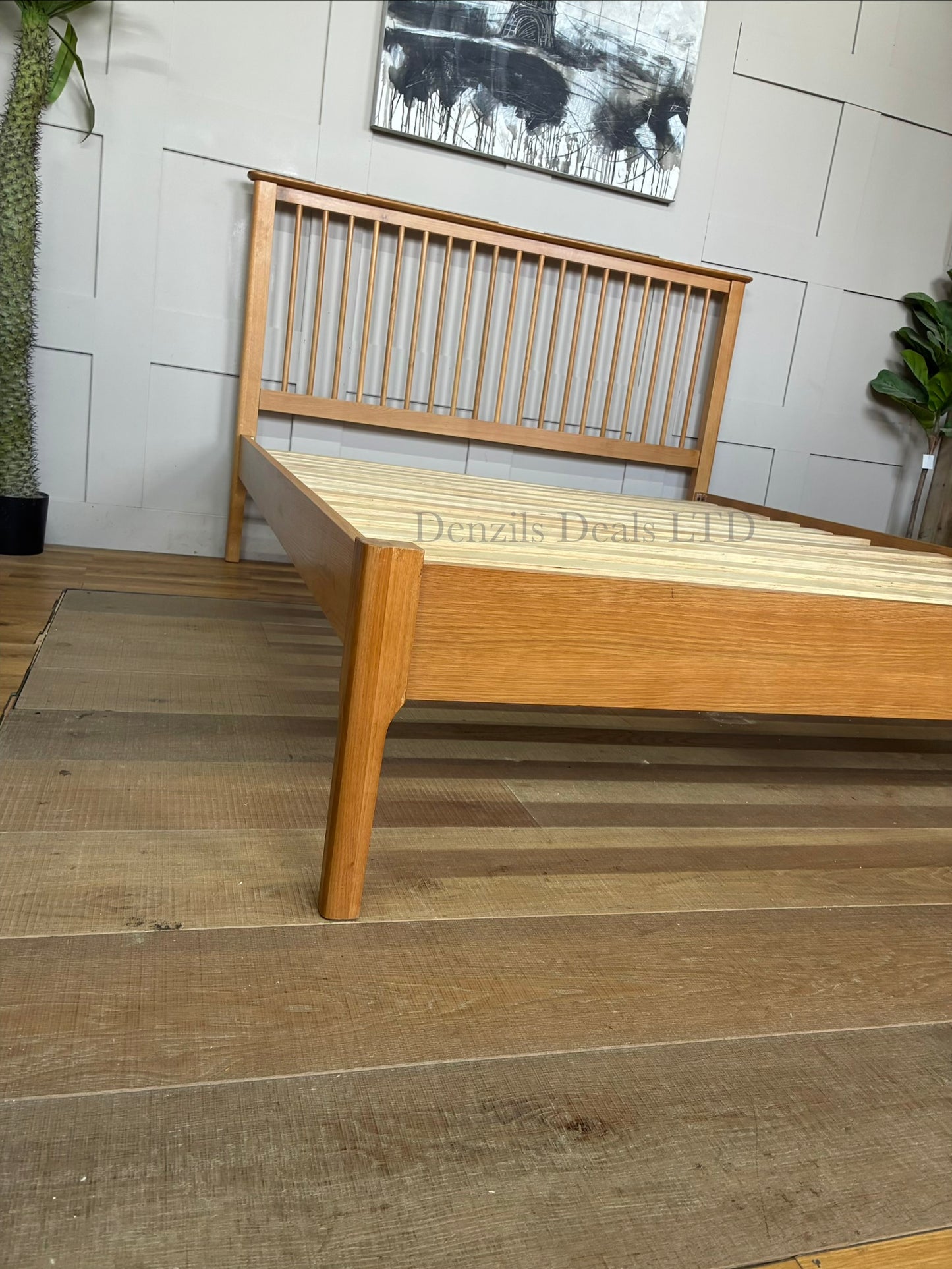 A king-size wooden bed frame made from light natural oak with a satin lacquer finish. The bed has a high headboard with vertical Shaker-style wooden slats, a low-profile matching footboard, and visible natural wood grain patterns