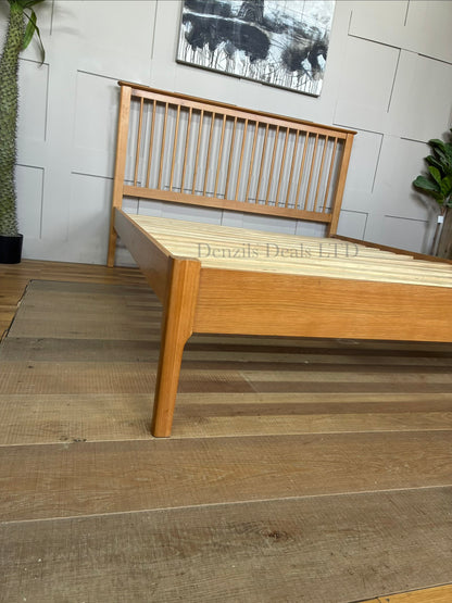 A king-size wooden bed frame made from light natural oak with a satin lacquer finish. The bed has a high headboard with vertical Shaker-style wooden slats, a low-profile matching footboard, and visible natural wood grain patterns