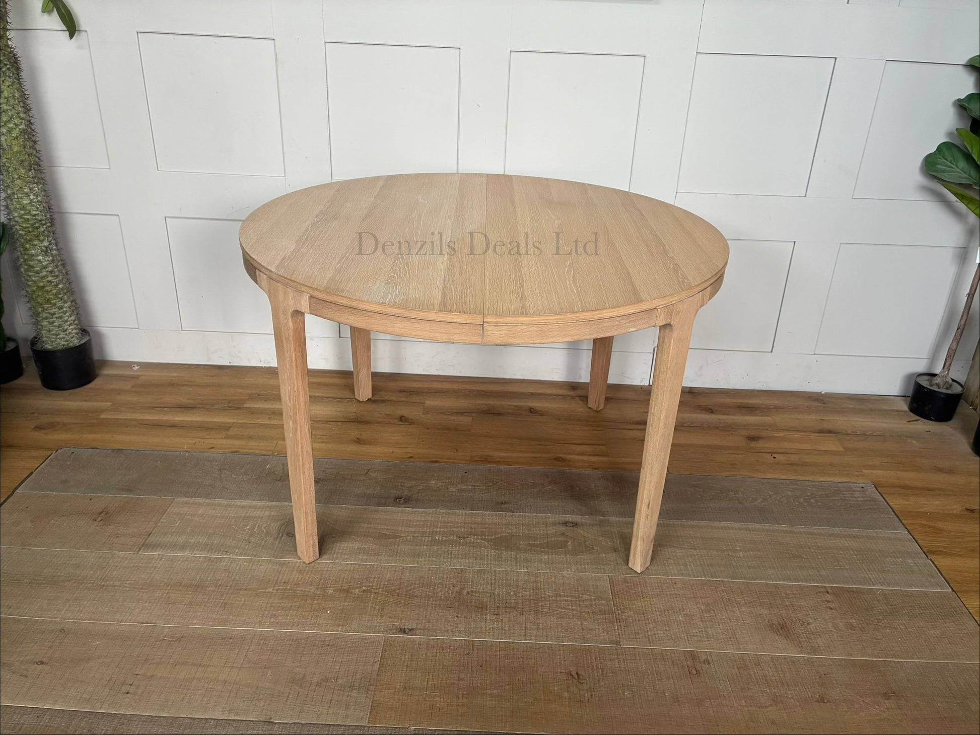A modern Scandi-style round dining table crafted from light natural oak with a prominent wood grain finish. The table features tapered, angled legs and an extending split-top design. It is shown in a bright, minimalist dining room setting