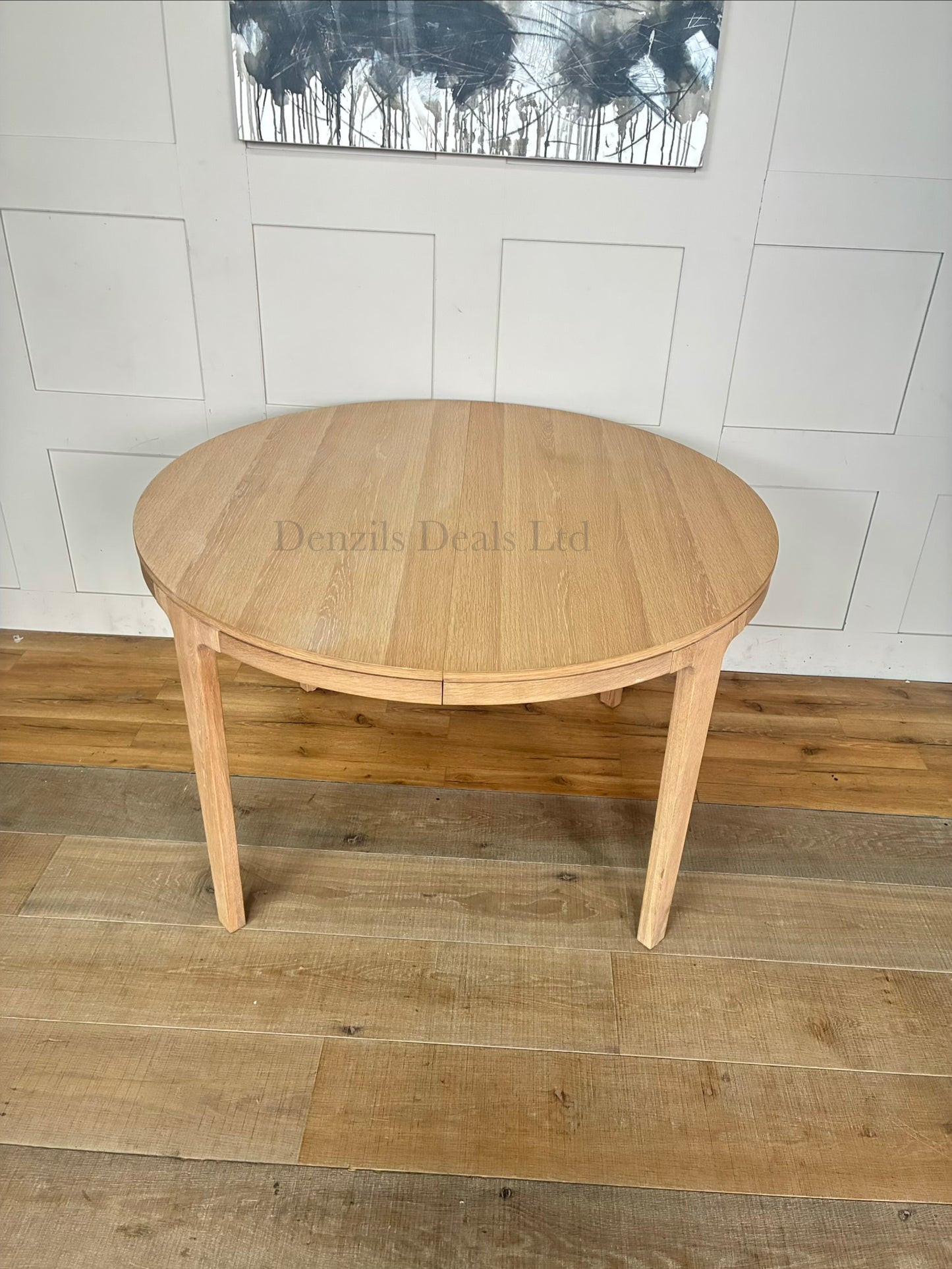 A modern Scandi-style round dining table crafted from light natural oak with a prominent wood grain finish. The table features tapered, angled legs and an extending split-top design. It is shown in a bright, minimalist dining room setting