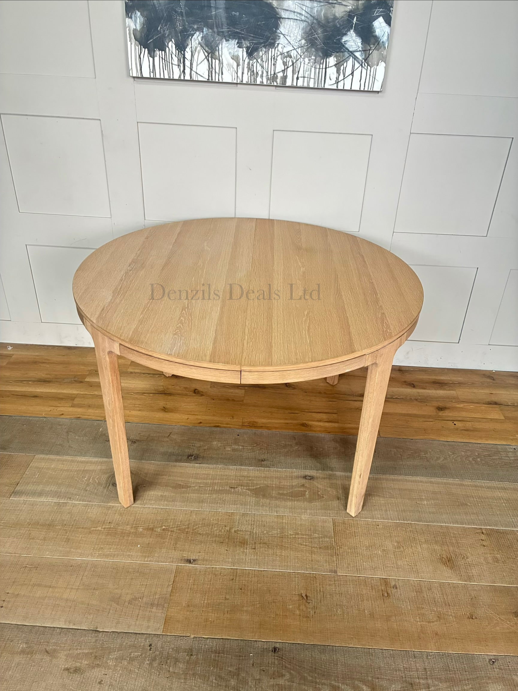 A modern Scandi-style round dining table crafted from light natural oak with a prominent wood grain finish. The table features tapered, angled legs and an extending split-top design. It is shown in a bright, minimalist dining room setting