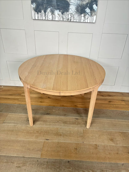 A modern Scandi-style round dining table crafted from light natural oak with a prominent wood grain finish. The table features tapered, angled legs and an extending split-top design. It is shown in a bright, minimalist dining room setting