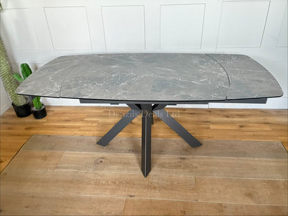 A modern rectangular extending dining table featuring a sleek printed glass top with an HPL finish and a smooth twist-to-open central extension mechanism, supported by a contemporary matte metal base."
