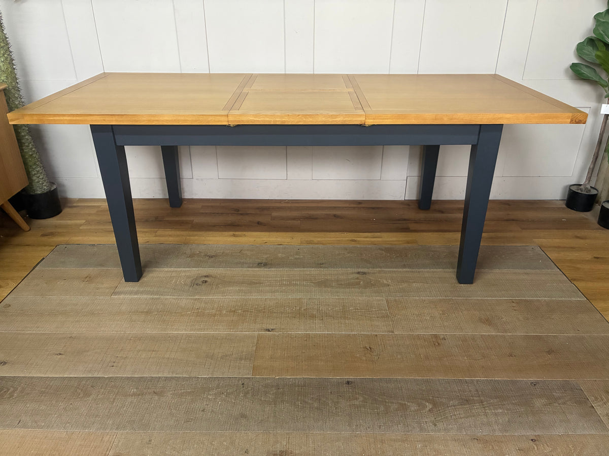 A rectangular wooden extending dining table from the DFS Country Living Maud range. The table features a premium navy blue painted finish on its sturdy legs and frame, contrasted by a smooth, natural oak wood grain tabletop with subtly rounded corners