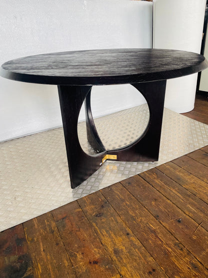 A round Anthropologie Iris dining table in a matte black oak finish, featuring a sculptural, curved pedestal base accented with gold-toned brass hardware