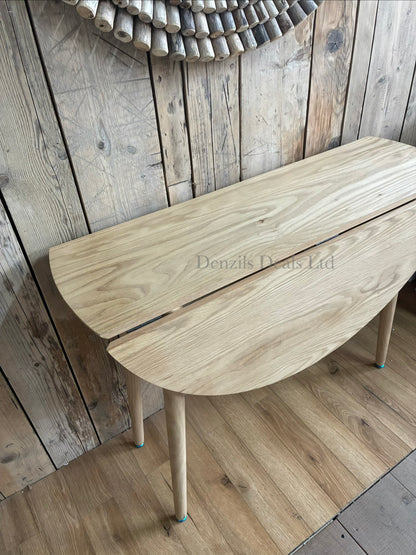 A round, space-saving wooden dining table with a smooth, light-toned natural oak veneer finish. The table is supported by a light wood frame featuring hinged gatelegs that swing out to hold up the folding drop leaves.