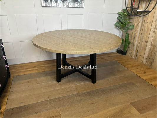 A round wooden dining table with a natural pine finish showing a subtle grain and patina, supported by sleek black metal or painted wooden legs that cross in the center.