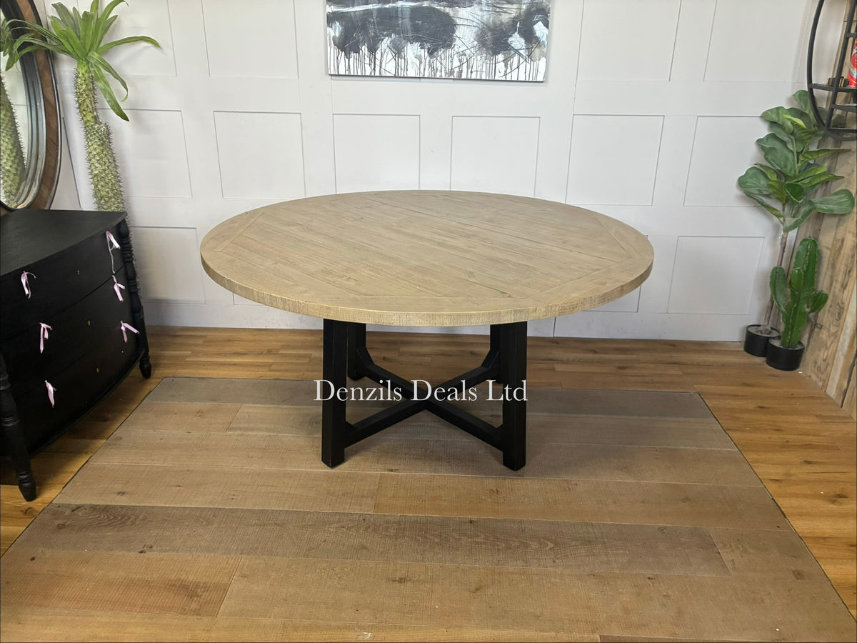 A round wooden dining table with a natural pine finish showing a subtle grain and patina, supported by sleek black metal or painted wooden legs that cross in the center.
