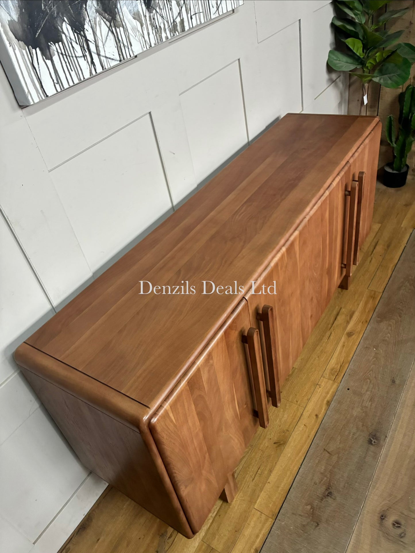 A side-facing product shot of the John Lewis Akola Sideboard in a light, natural wood tone. The sideboard is crafted from panelled acacia wood, creating vertical line patterns across its four minimalist doors. The outer edges of the frame are softly rounded, softening its blocky silhouette. The unit sits directly on the ground without legs, serving as a clean, mid-century or modern living room storage solution