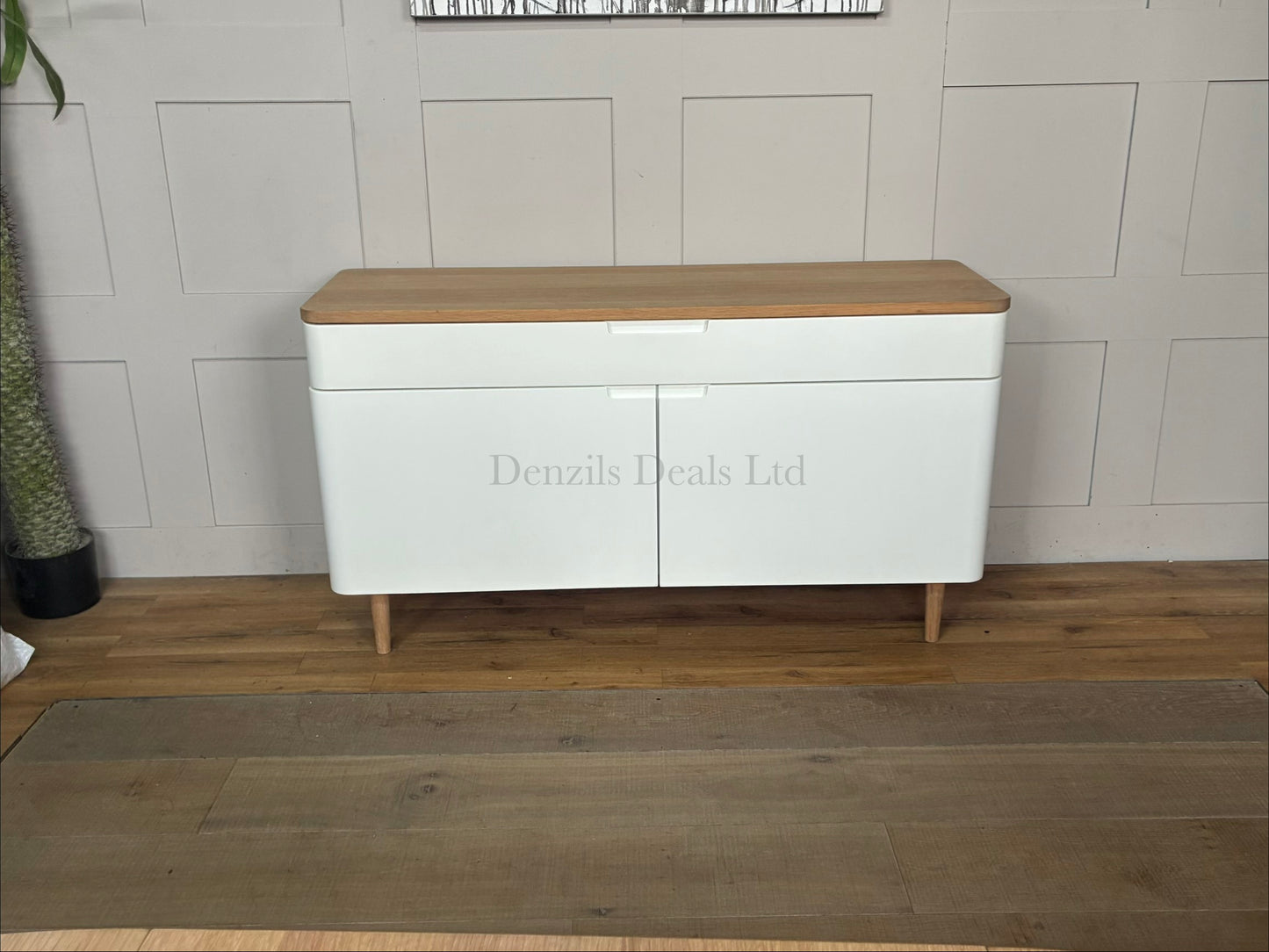 A wide Scandinavian-style sideboard featuring a white painted frame and cabinet doors, a natural solid oak top, a wide divided top drawer with integrated handles, smooth rounded corners, and sleek tapered wooden legs.