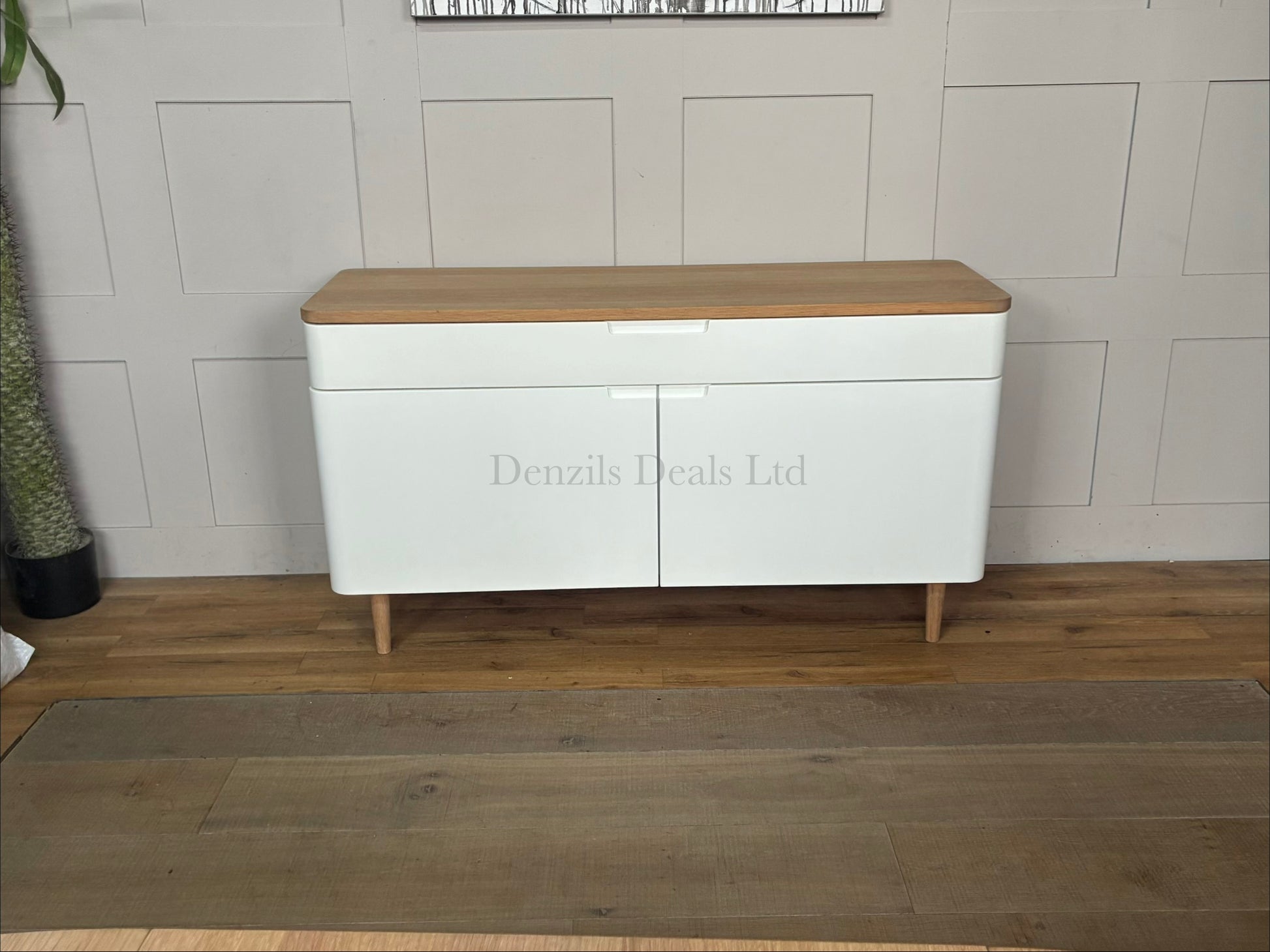 A wide Scandinavian-style sideboard featuring a white painted frame and cabinet doors, a natural solid oak top, a wide divided top drawer with integrated handles, smooth rounded corners, and sleek tapered wooden legs.
