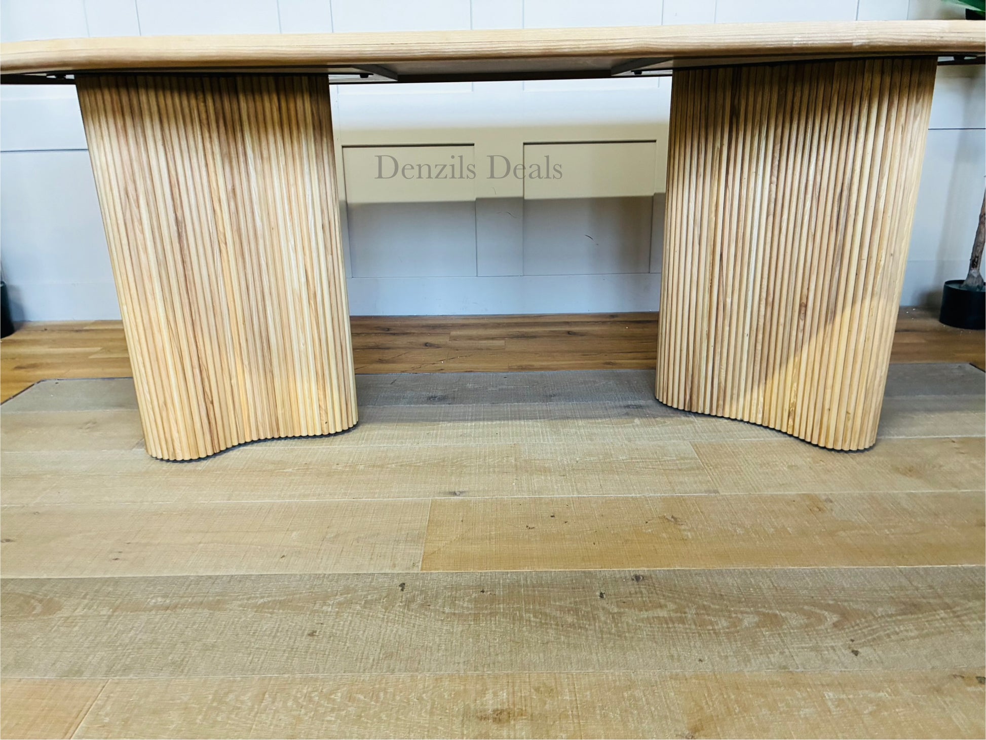 An Anthropologie Scout oval dining table made of light beige ash wood, featuring an oval tabletop with a decorative burl veneer pattern and thick, fluted pedestal legs. 