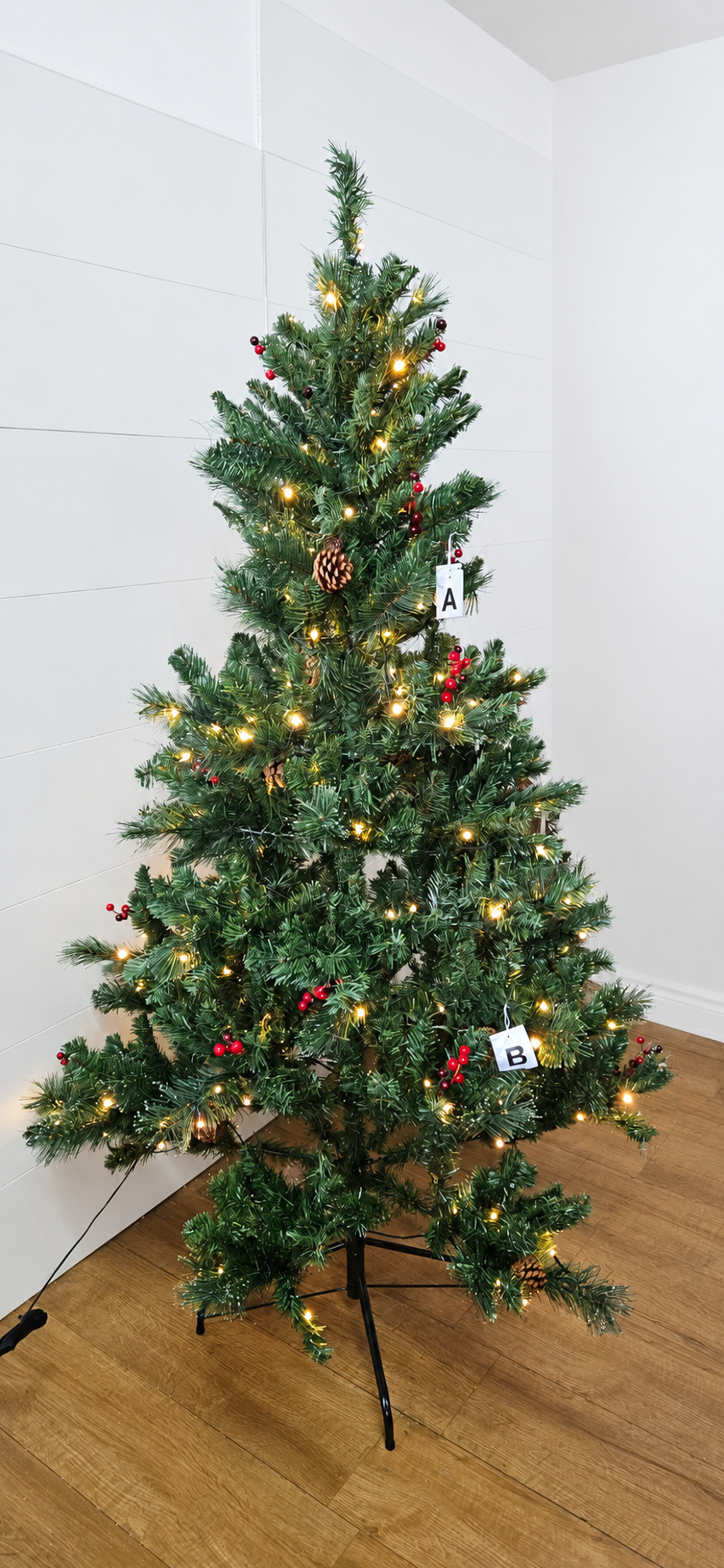 An M&S six-foot green pine-style artificial Christmas tree. The tree features realistic green needles decorated with clusters of bright red berries and natural pine cones, pre-lit with 200 warm white LED lights that emit a soft holiday glow