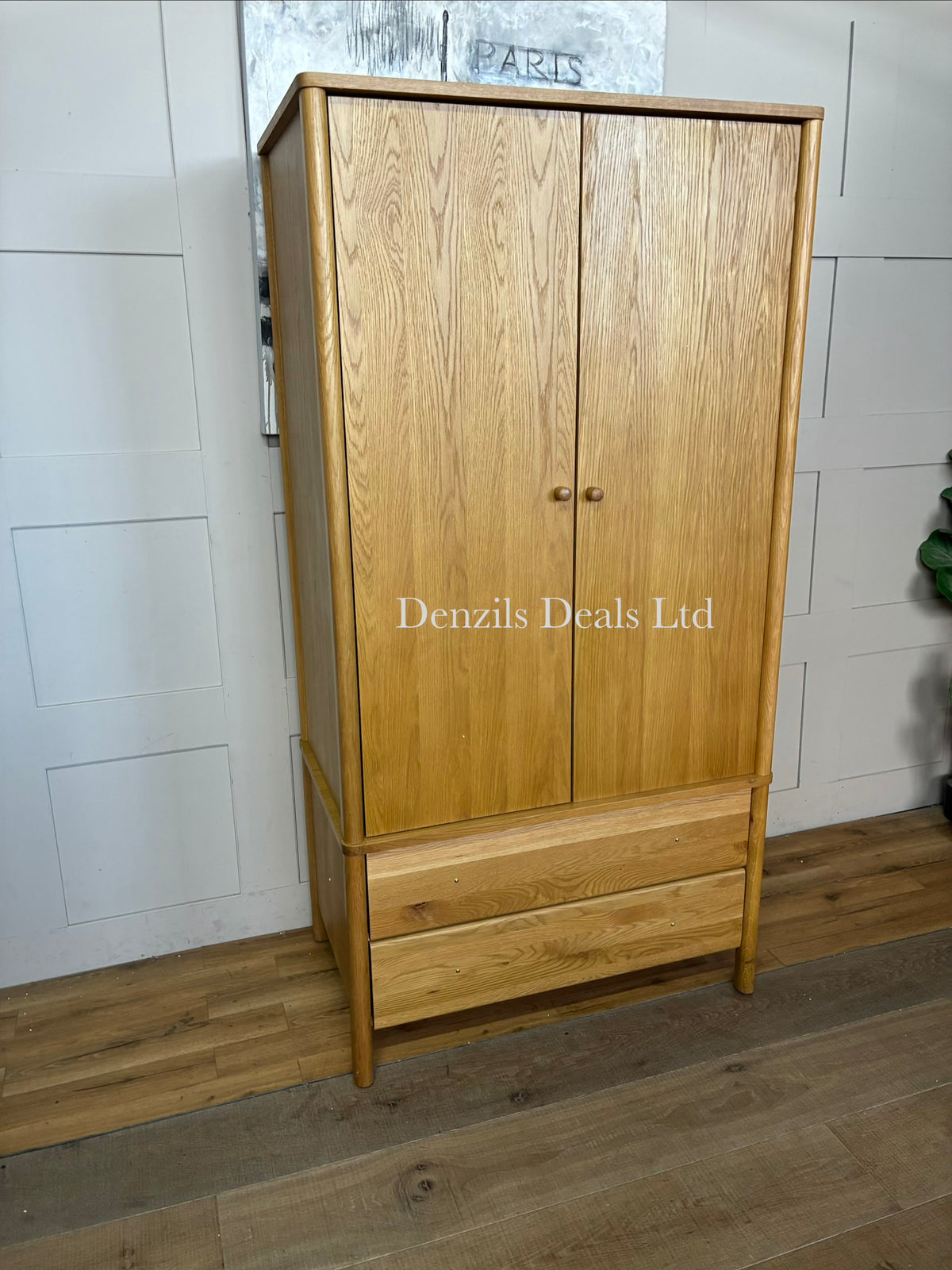 An elegant John Lewis, mid-century modern freestanding double wardrobe crafted from warm, natural oak. The piece features two large doors with softer, rounded edges and clean minimalist vertical lines, opening to reveal a wooden hanging rail inside. Beneath the main cabinet section sit two wide, deep drawers stacked vertically for additional storage. The entire unit is accented by small, solid turned timber handles on both the doors and drawer fronts, giving it a smooth, unified, and contemporary look