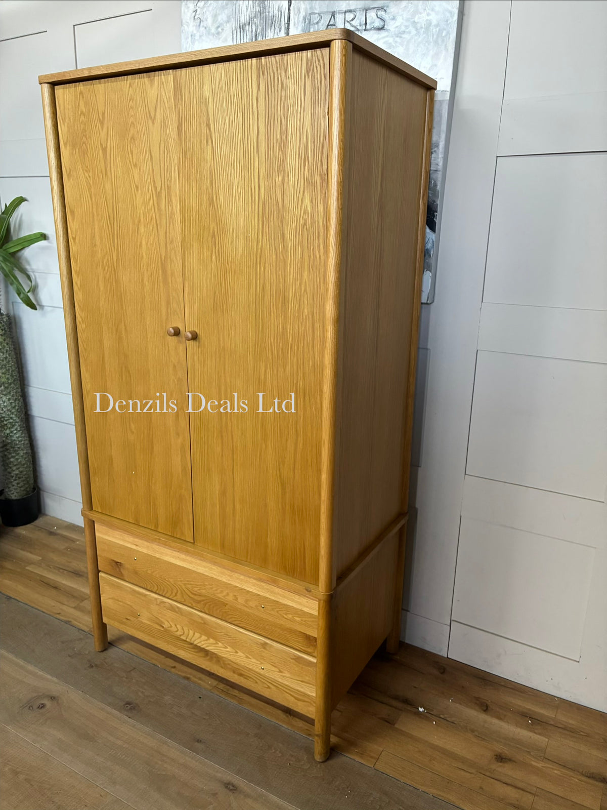 An elegant John Lewis, mid-century modern freestanding double wardrobe crafted from warm, natural oak. The piece features two large doors with softer, rounded edges and clean minimalist vertical lines, opening to reveal a wooden hanging rail inside. Beneath the main cabinet section sit two wide, deep drawers stacked vertically for additional storage. The entire unit is accented by small, solid turned timber handles on both the doors and drawer fronts, giving it a smooth, unified, and contemporary look
