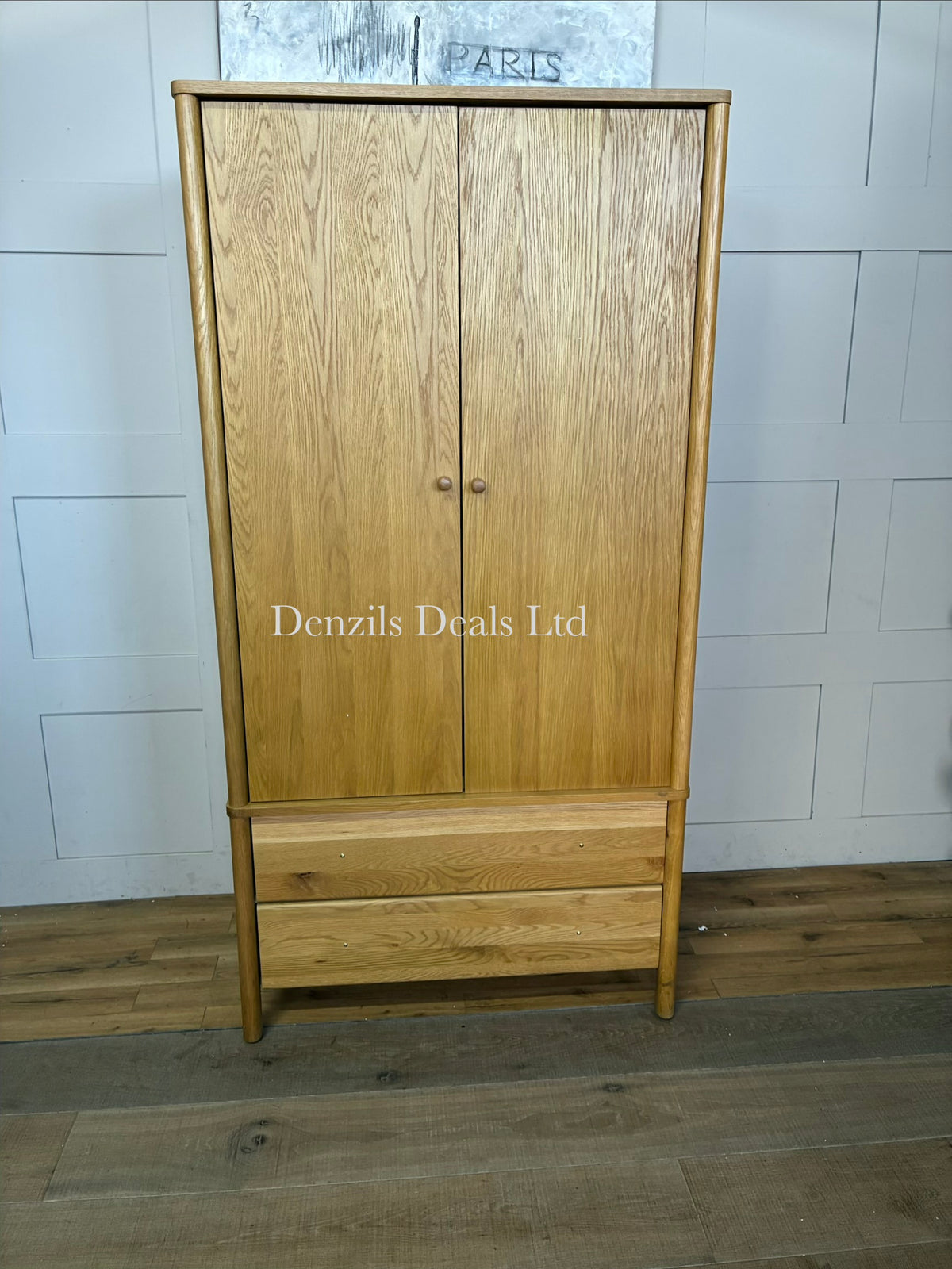 An elegant, mid-century modern freestanding double wardrobe crafted from warm, natural oak. The piece features two large doors with softer, rounded edges and clean minimalist vertical lines, opening to reveal a wooden hanging rail inside. Beneath the main cabinet section sit two wide, deep drawers stacked vertically for additional storage. The entire unit is accented by small, solid turned timber handles on both the doors and drawer fronts, giving it a smooth, unified, and contemporary look