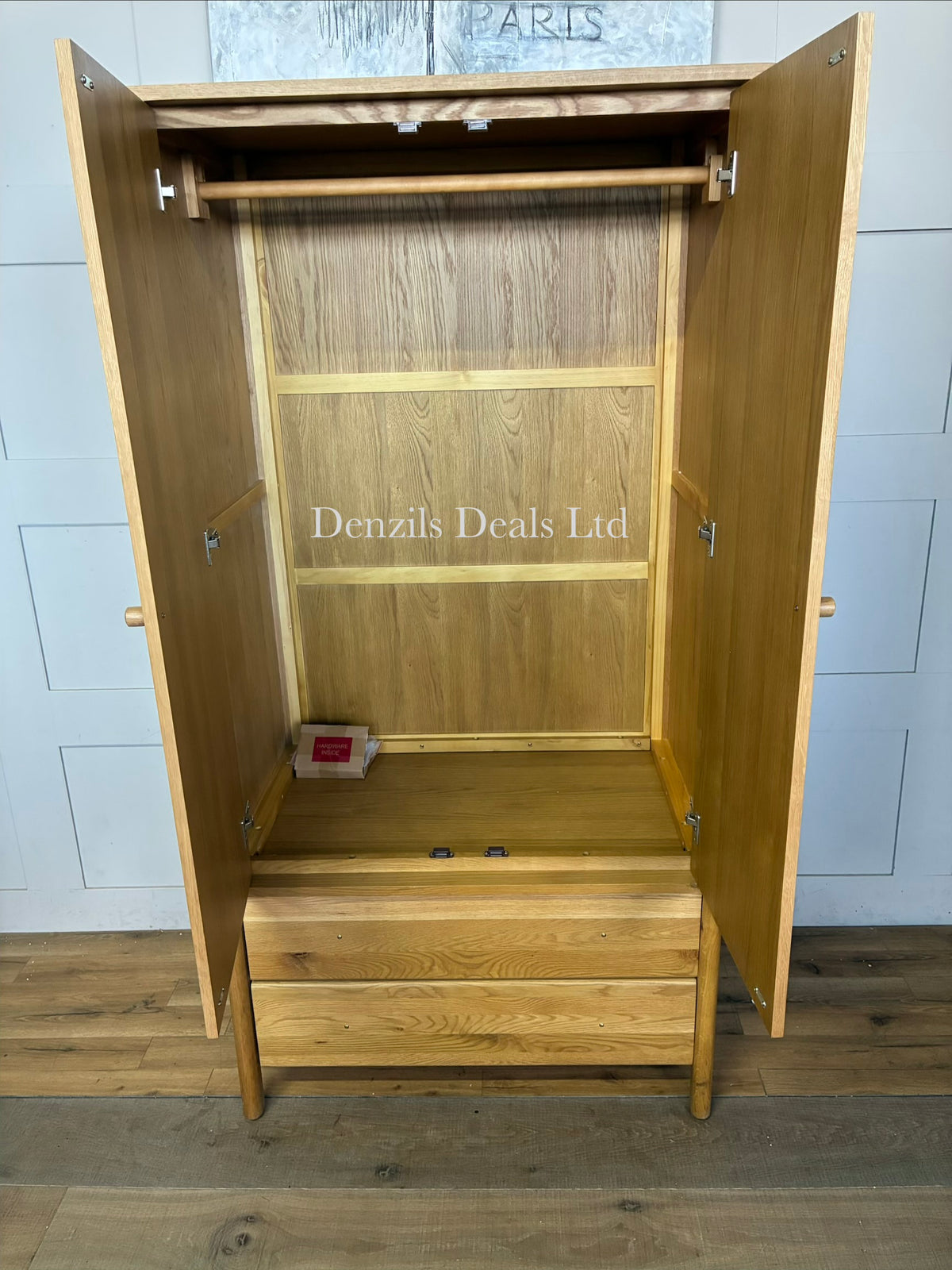 An elegant John Lewis, mid-century modern freestanding double wardrobe crafted from warm, natural oak. The piece features two large doors with softer, rounded edges and clean minimalist vertical lines, opening to reveal a wooden hanging rail inside. Beneath the main cabinet section sit two wide, deep drawers stacked vertically for additional storage. The entire unit is accented by small, solid turned timber handles on both the doors and drawer fronts, giving it a smooth, unified, and contemporary look