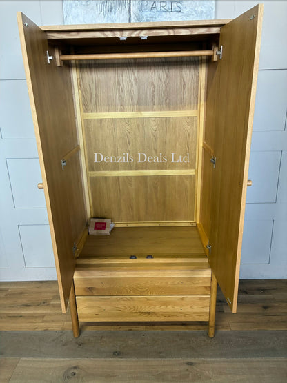An elegant John Lewis, mid-century modern freestanding double wardrobe crafted from warm, natural oak. The piece features two large doors with softer, rounded edges and clean minimalist vertical lines, opening to reveal a wooden hanging rail inside. Beneath the main cabinet section sit two wide, deep drawers stacked vertically for additional storage. The entire unit is accented by small, solid turned timber handles on both the doors and drawer fronts, giving it a smooth, unified, and contemporary look