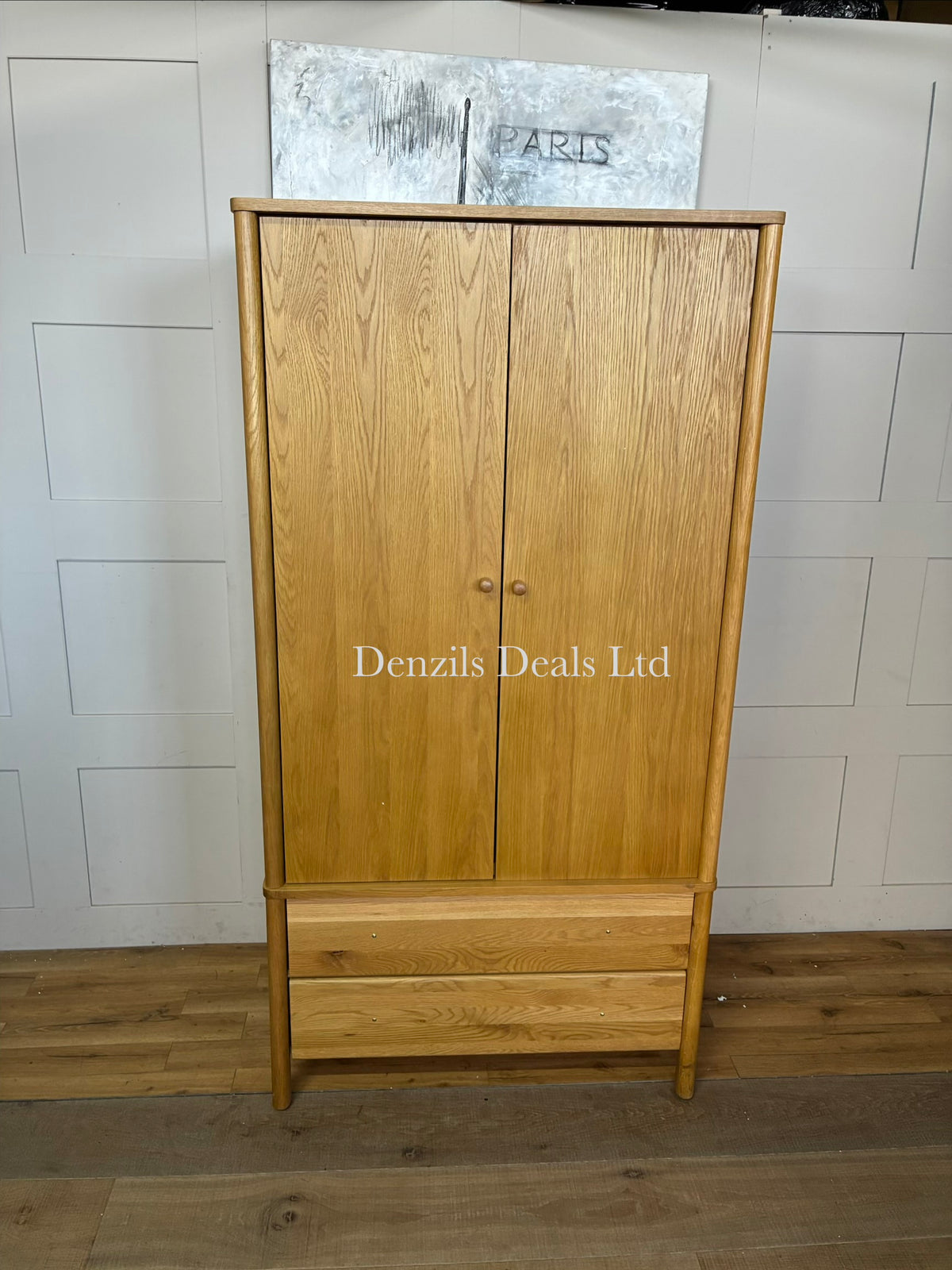 An elegant, mid-century modern freestanding double wardrobe crafted from warm, natural oak. The piece features two large doors with softer, rounded edges and clean minimalist vertical lines, opening to reveal a wooden hanging rail inside. Beneath the main cabinet section sit two wide, deep drawers stacked vertically for additional storage. The entire unit is accented by small, solid turned timber handles on both the doors and drawer fronts, giving it a smooth, unified, and contemporary look